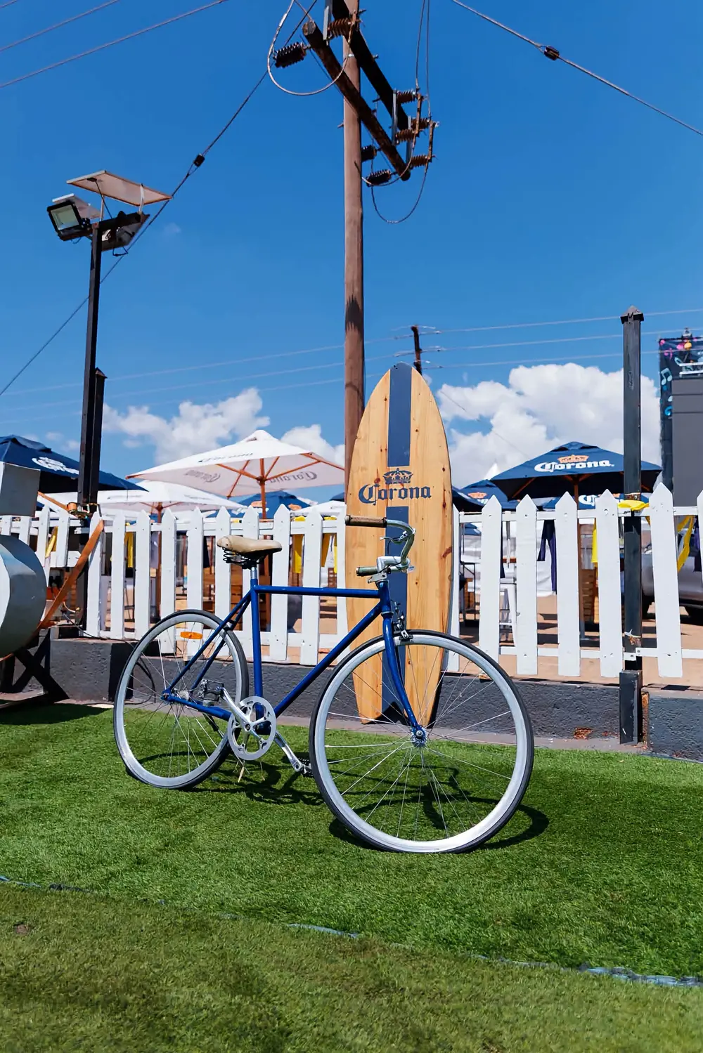 Blue vintage bicycle and Corona surfboard against a white picket fence